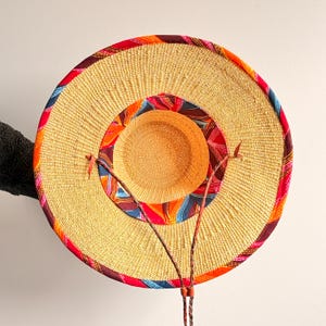 Puede incluir: Un sombrero de paja de ala ancha con una textura tejida en tono beige natural. El sombrero presenta un borde de tela multicolor en tonos rojos, naranjas y azules a lo largo del ala exterior y la corona interior, con un cord&oacute;n trenzado fino.