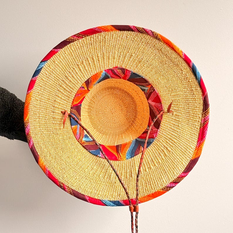Puede incluir: Un sombrero de paja de ala ancha con una textura tejida en tono beige natural. El sombrero presenta un borde de tela multicolor en tonos rojos, naranjas y azules a lo largo del ala exterior y la corona interior, con un cord&oacute;n trenzado fino.