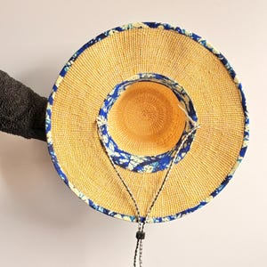 Puede incluir: Un sombrero de paja de ala ancha con una textura tejida en tono bronceado natural. El sombrero est&aacute; acentuado con un borde de tela con estampado azul y blanco a lo largo del borde exterior y la corona interior. Incluye una correa ajustable.