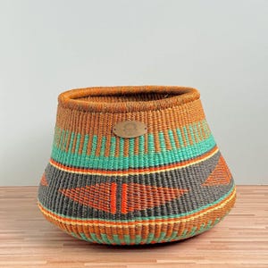 May include: A woven basket featuring a wide, rounded base and a tapered opening. The exterior displays horizontal bands of orange, teal, and charcoal grey, accented by geometric diamond patterns in orange. An oval leather tag is centred near the rim.