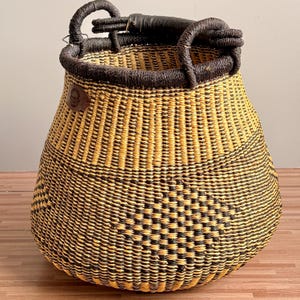 May include: A round, woven basket featuring a geometric pattern in alternating black and natural yellow straw. The basket has a dark, thick, wrapped handle at the top and a small, circular brown leather patch with an embossed logo on the side.