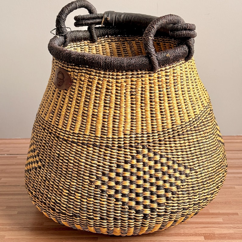 May include: A round, woven basket featuring a geometric pattern in alternating black and natural yellow straw. The basket has a dark, thick, wrapped handle at the top and a small, circular brown leather patch with an embossed logo on the side.