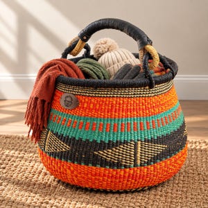 May include: A round, hand-woven basket featuring horizontal bands of orange, black, and teal with geometric triangle patterns. It has a black wrapped handle and a leather patch. The basket is filled with a rust-colored scarf, a beige pom-pom hat, and green yarn.