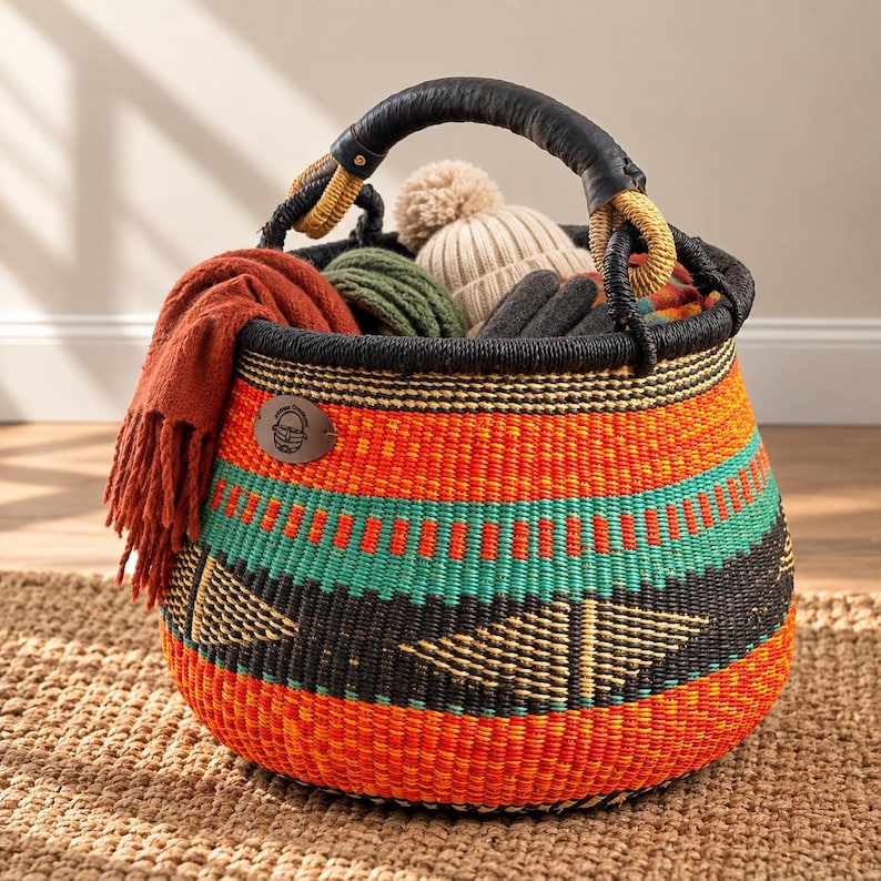 May include: A round, hand-woven basket featuring horizontal bands of orange, black, and teal with geometric triangle patterns. It has a black wrapped handle and a leather patch. The basket is filled with a rust-colored scarf, a beige pom-pom hat, and green yarn.