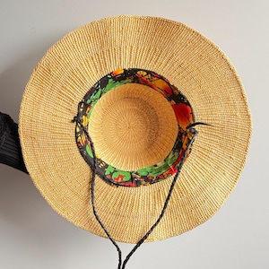 Puede incluir: Un sombrero de paja de ala ancha con una textura tejida en color beige natural. El interior de la copa presenta un forro de tela colorido con un patr&oacute;n abstracto en negro, rojo, amarillo y verde. Incluye una fina correa de barbilla de cord&oacute;n negro trenzado.