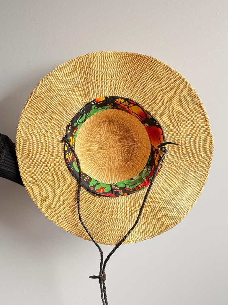 Puede incluir: Un sombrero de paja de ala ancha con una textura tejida en color beige natural. El interior de la copa presenta un forro de tela colorido con un patr&oacute;n abstracto en negro, rojo, amarillo y verde. Incluye una fina correa de barbilla de cord&oacute;n negro trenzado.