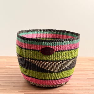 May include: A round, hand-woven basket featuring horizontal stripes in vibrant shades of hot pink, lime green, and black. The basket has a wide, open top and a dark brown, oval-shaped leather patch centred on the front exterior surface.