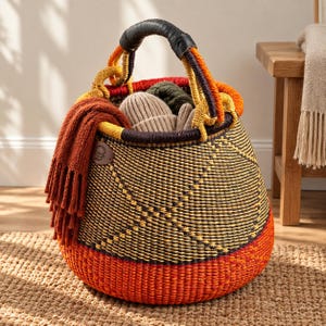 May include: A round woven basket featuring a black and yellow geometric pattern with a solid orange base. The basket has a thick, dark handle and contains a rust-colored knit scarf and several neutral-toned knit hats tucked inside the rim.