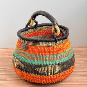 May include: A round woven basket featuring horizontal bands of bright orange, teal, and dark brown. The basket has a thick, dark brown wrapped handle and a small, circular brown leather-like patch on the side with an embossed basket icon.