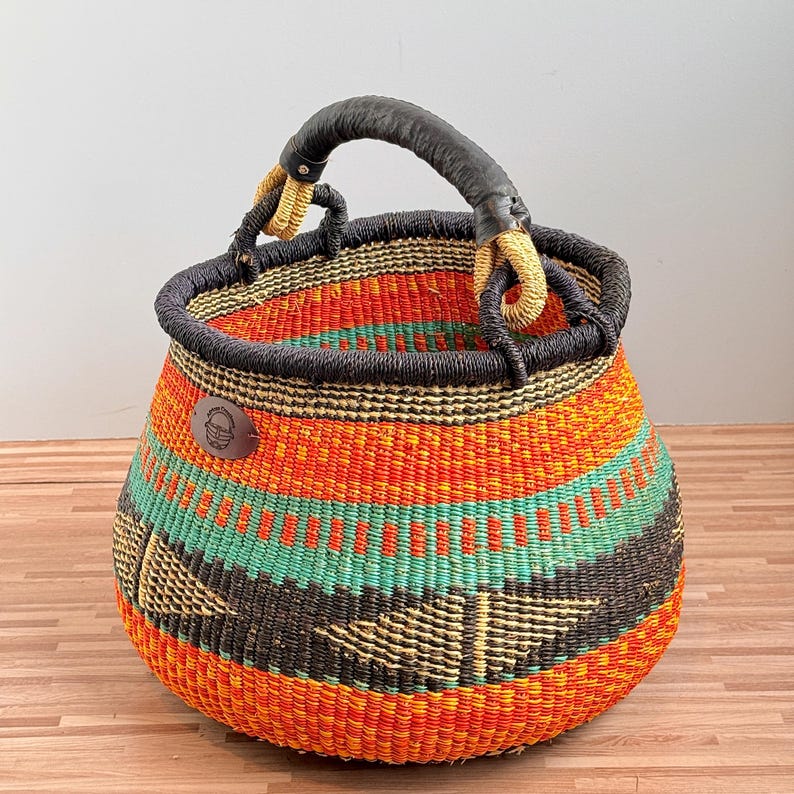 May include: A round woven basket featuring horizontal bands of bright orange, teal, and dark brown. The basket has a thick, dark brown wrapped handle and a small, circular brown leather-like patch on the side with an embossed basket icon.