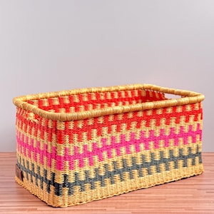 May include: A rectangular woven basket featuring a horizontal striped pattern in vibrant red, bright pink, and dark grey, contrasted with natural tan fibres. The basket has a sturdy, reinforced rim and a small brown rectangular tag on the side.