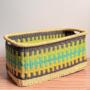 May include: A rectangular woven storage basket featuring a repeating zigzag pattern in vibrant shades of lime green, turquoise, and yellow, contrasted with dark charcoal grey. The basket has a natural tan rim and a matching woven base.