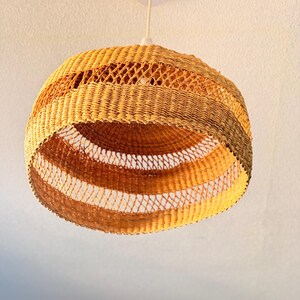 K&ouml;nnte beinhalten: Eine gewebte Pendelleuchte mit kugelf&ouml;rmiger Form. Die Leuchte besteht aus naturfarbenem Korbgeflecht und hat ein geschichtetes Design mit hell- und dunkelbraunen B&auml;ndern. Das offene Gewebe l&auml;sst Licht durchscheinen.