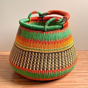 May include: A round, hand-woven basket featuring horizontal bands of vibrant green, bright orange, yellow, and dark brown. The basket has a wide, open top with a thick, wrapped handle in a reddish-brown tone and a small circular leather logo patch.