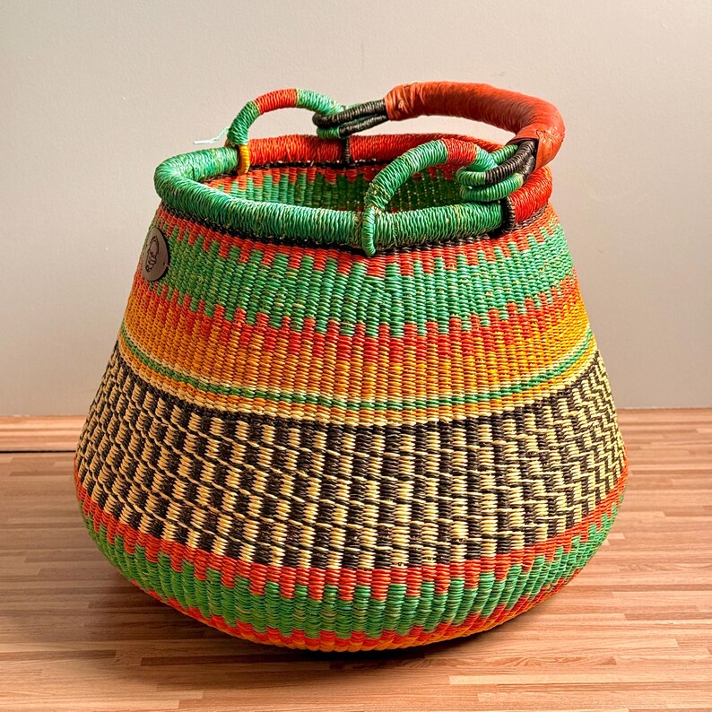 May include: A round, hand-woven basket featuring horizontal bands of vibrant green, bright orange, yellow, and dark brown. The basket has a wide, open top with a thick, wrapped handle in a reddish-brown tone and a small circular leather logo patch.