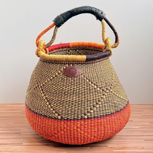 May include: A round woven basket featuring a vibrant orange base, a body with a yellow and dark brown geometric pattern, and a thick black leather-wrapped handle. An oval brown leather patch with a basket icon is centered on the upper front.