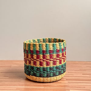 Può includere: Un cesto cilindrico intrecciato con un motivo geometrico colorato. Il cesto presenta fasce orizzontali gialle, verdi, rosse e nere. Il cesto è realizzato con fibre naturali e poggia su una superficie in legno.