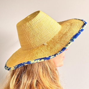 Puede incluir: Un sombrero de paja de ala ancha con una textura tejida en color tostado natural y una copa plana. El borde exterior del ala est&aacute; rematado con un ribete de tela azul vibrante con un estampado floral blanco y amarillo que contrasta.