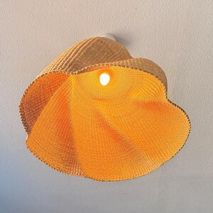 K&ouml;nnte beinhalten: Eine gewebte Pendelleuchte aus Rattan. Die Leuchte hat eine warme, nat&uuml;rliche, hellbraune Farbe und eine bl&uuml;tenartige Form. Das Innere der Leuchte wird von einer einzelnen Gl&uuml;hbirne beleuchtet, die ein warmes orangefarbenes Leuchten erzeugt. Die Leuchte ist an einer wei&szlig;en Decke montiert.