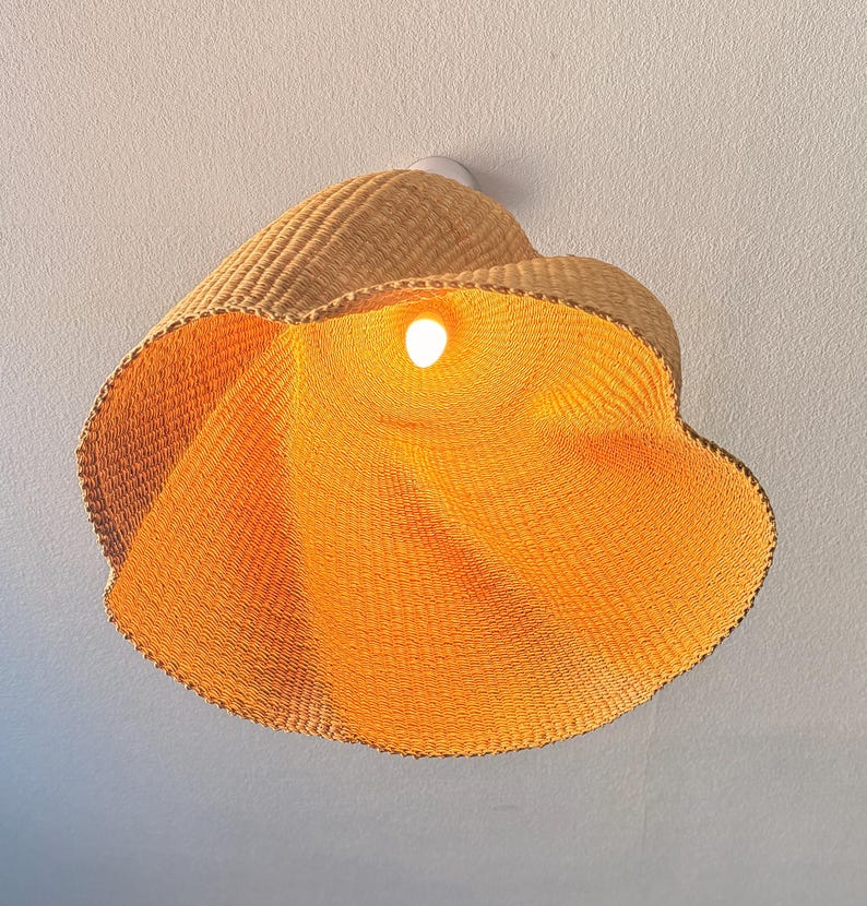 K&ouml;nnte beinhalten: Eine gewebte Pendelleuchte aus Rattan. Die Leuchte hat eine warme, nat&uuml;rliche, hellbraune Farbe und eine bl&uuml;tenartige Form. Das Innere der Leuchte wird von einer einzelnen Gl&uuml;hbirne beleuchtet, die ein warmes orangefarbenes Leuchten erzeugt. Die Leuchte ist an einer wei&szlig;en Decke montiert.