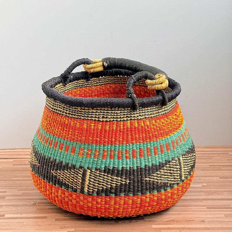 May include: A round woven basket featuring horizontal bands of bright orange, teal, and black. The basket has a thick, dark-colored rim and two sturdy handles wrapped in light-colored cord, with a smooth black leather-like grip on the top handle.