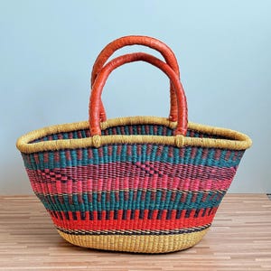 May include: A woven basket with two orange leather-wrapped handles. The body features horizontal bands of yellow, teal, pink, and red woven fibres in a repeating pattern. The rim is finished with a thick, yellow woven border.