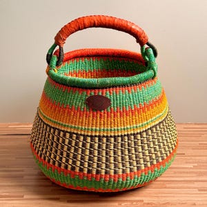 May include: A round woven basket featuring a vibrant pattern of horizontal stripes in bright orange, yellow, and green, with a dark brown checkered band near the base. It has a sturdy orange wrapped handle and a small brown oval leather-like patch.