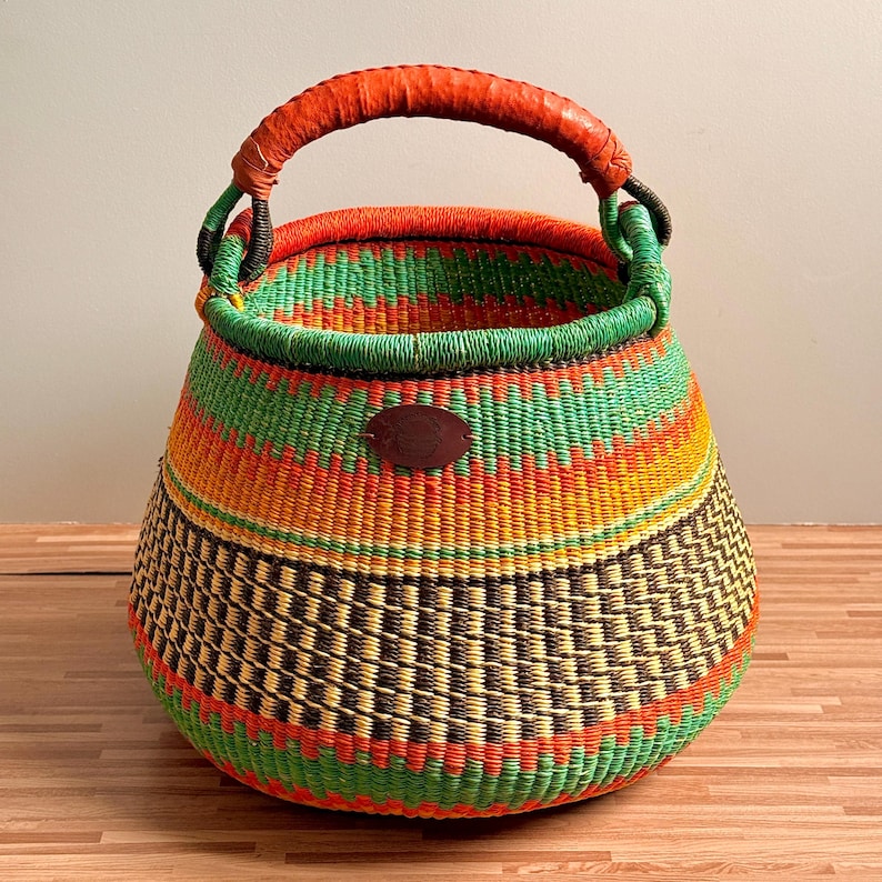May include: A round woven basket featuring a vibrant pattern of horizontal stripes in bright orange, yellow, and green, with a dark brown checkered band near the base. It has a sturdy orange wrapped handle and a small brown oval leather-like patch.