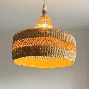 K&ouml;nnte beinhalten: Eine Pendelleuchte aus geflochtenem Rattan. Die Lampe hat einen zylindrischen Schirm mit einem gewebten Muster in Braun- und Beiget&ouml;nen mit einem orangefarbenen Band. Das Licht ist an und erzeugt einen warmen Schein. Die Leuchte h&auml;ngt an einem wei&szlig;en Kabel.