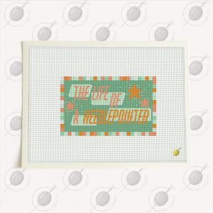 May include: A needlepoint canvas with the text "THE LIFE OF A NEEDLEPOINTER" in orange and white on a green background. The canvas is framed in white and includes orange stars and pink accents.