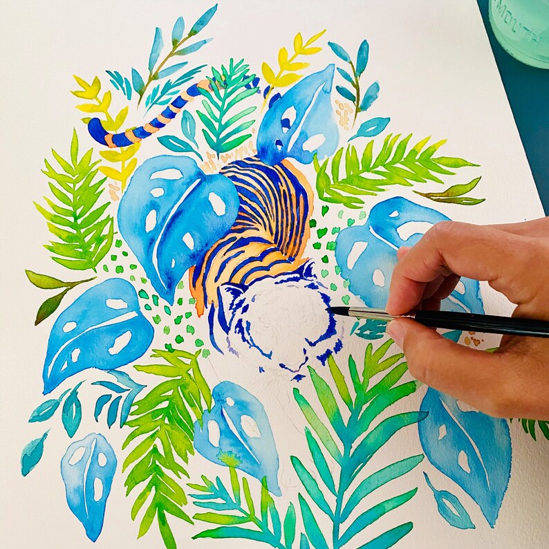 Puede incluir: Pintura acuarela de un tigre con follaje azul y verde. El tigre est&aacute; acostado con la cabeza girada hacia un lado. El tigre est&aacute; pintado en tonos de naranja, amarillo y blanco. El follaje est&aacute; pintado en tonos de azul, verde y amarillo.