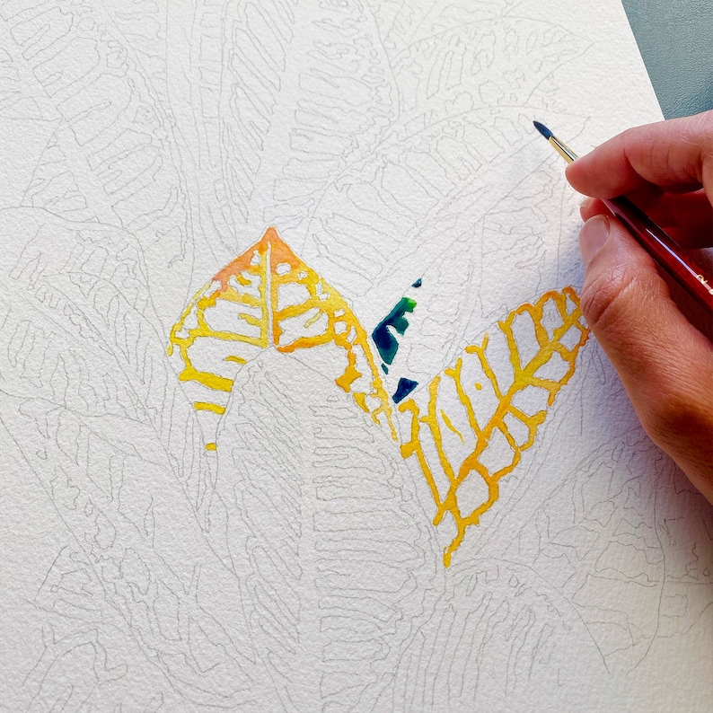 Puede incluir: Un primer plano de una pintura de acuarela en proceso. El artista est&aacute; usando un pincel para agregar pintura amarilla y naranja a un dise&ntilde;o de hoja. La hoja est&aacute; delineada en negro y el fondo es blanco.