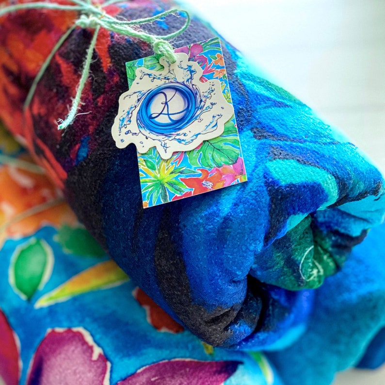 May include: Rolled beach towels with vibrant watercolor floral patterns in shades of blue, orange, and purple. A tag with a blue swirl design and the letter "K" is attached with green twine. The towels are soft and plush.