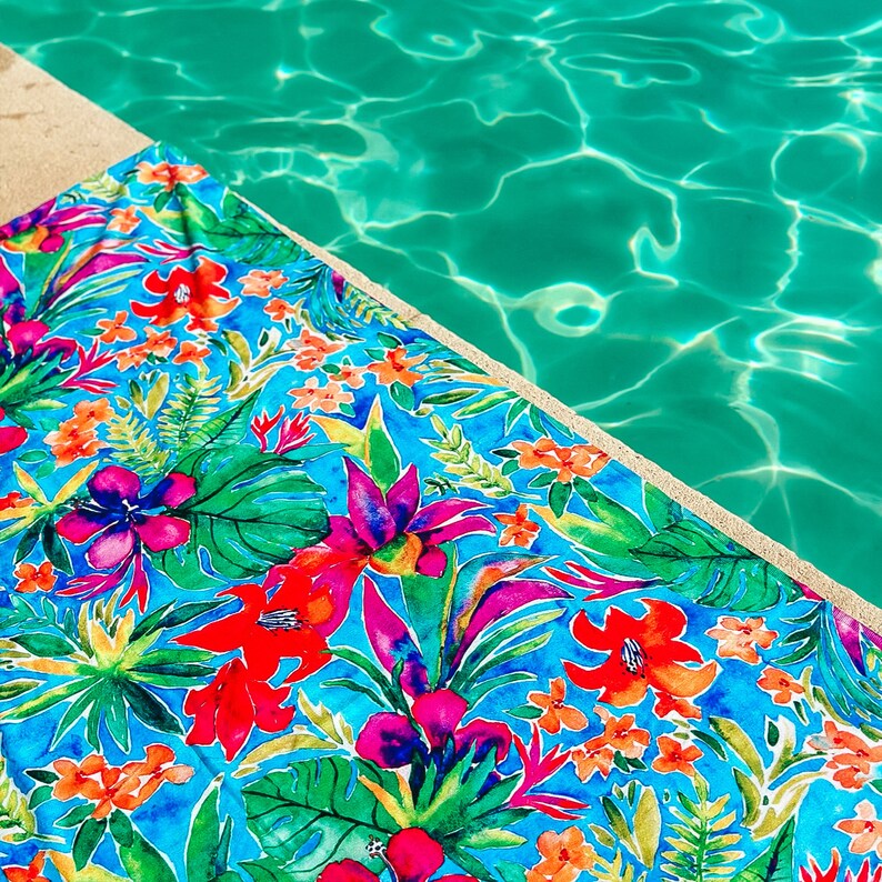 May include: A vibrant beach towel with a tropical floral pattern, featuring large red, pink, and orange flowers, and green leaves on a bright blue background. The towel is placed near a pool with clear turquoise water.