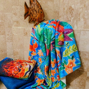 May include: A vibrant beach towel with a tropical floral design in shades of blue, green, orange, and purple. A second towel with a colorful abstract design is stacked on top of a blue towel. A wooden fish decorates the wall.