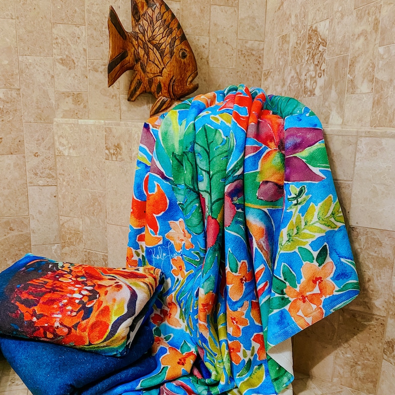 May include: A vibrant beach towel with a tropical floral design in shades of blue, green, orange, and purple. A second towel with a colorful abstract design is stacked on top of a blue towel. A wooden fish decorates the wall.
