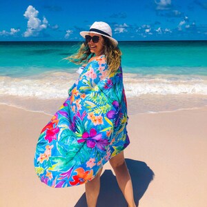 May include: A vibrant, flowing beach cover-up with a tropical floral print in shades of blue, pink, orange, and green. The cover-up is being worn on a sunny beach with turquoise water and a white hat.