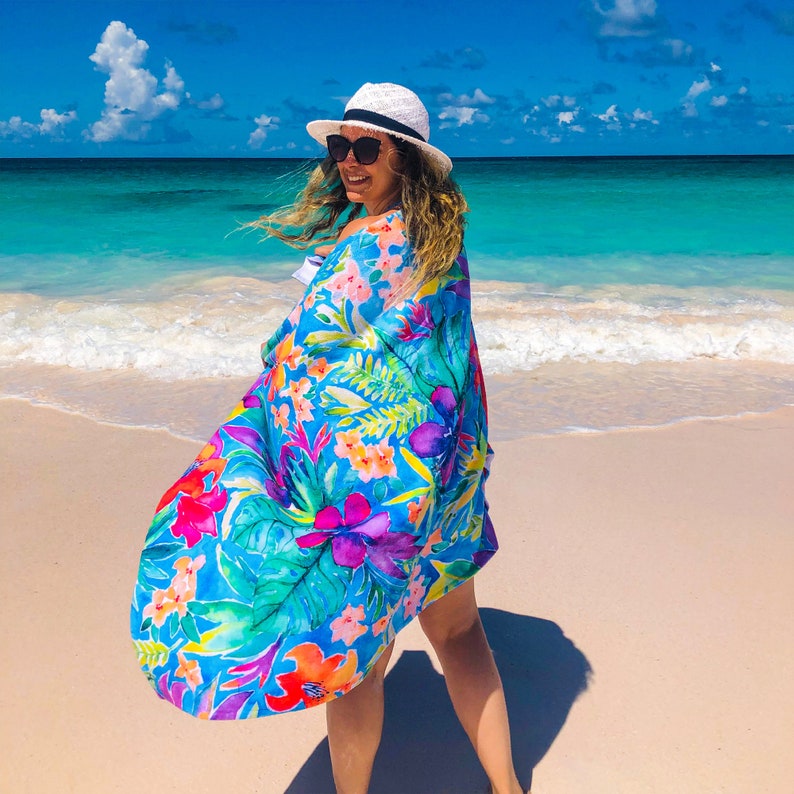 May include: A vibrant, flowing beach cover-up with a tropical floral print in shades of blue, pink, orange, and green. The cover-up is being worn on a sunny beach with turquoise water and a white hat.