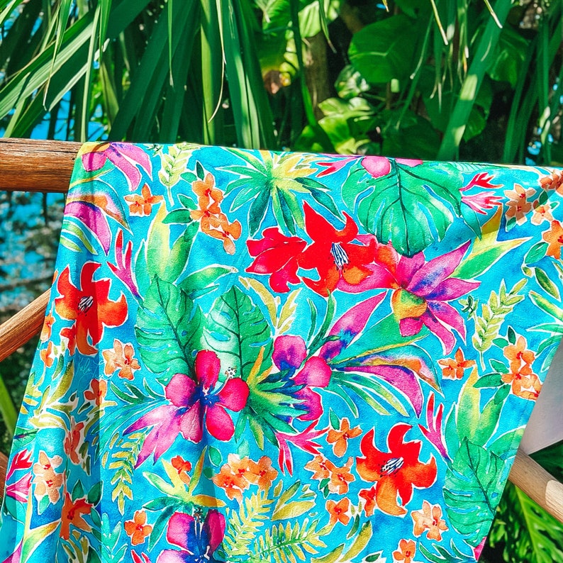 May include: A vibrant turquoise fabric with a tropical floral pattern. The design features large red, pink, and orange flowers, along with green leaves. The fabric is draped over a wooden railing, with lush green foliage in the background.