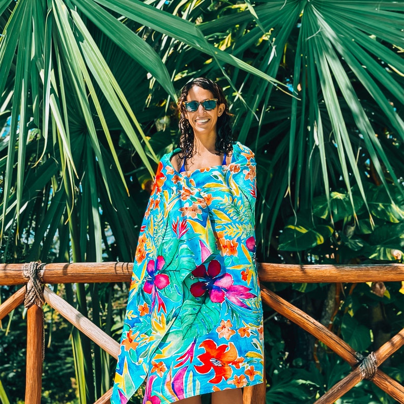 May include: A vibrant beach towel with a tropical floral pattern in shades of blue, green, red, and orange. The towel is wrapped around a person standing near a wooden railing and lush green foliage.
