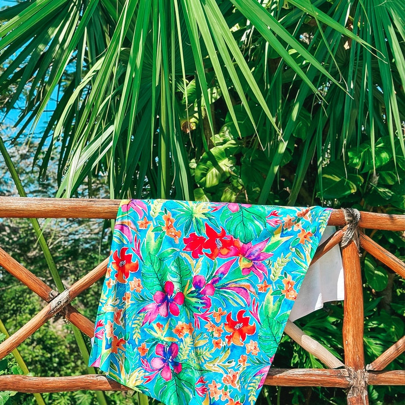 May include: A vibrant turquoise beach towel with a tropical floral pattern of red, pink, and purple flowers and green leaves. The towel is draped over a wooden railing, with lush green foliage in the background.