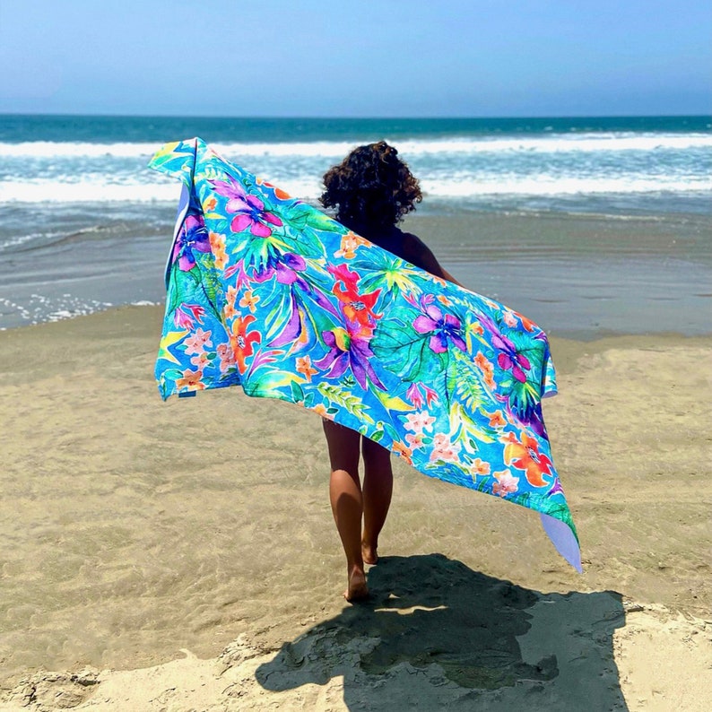 May include: A vibrant beach towel with a tropical floral pattern in shades of blue, green, pink, and orange is held up on a sandy beach. The towel is being held by a person walking towards the ocean. The ocean and sky are visible in the background.
