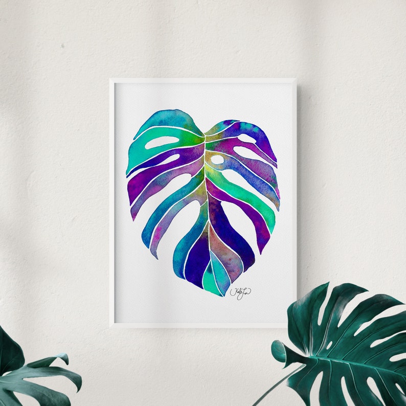 May include: A watercolor painting of a single monstera leaf in shades of blue, purple, and green. The leaf is framed in a white frame.