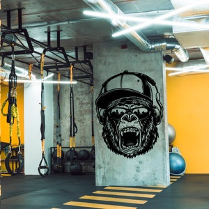 May include: A black and white illustration of a gorilla wearing a baseball cap and headphones, with a city skyline reflected in its sunglasses. The illustration is on a concrete wall in a gym with yellow walls and a black floor.