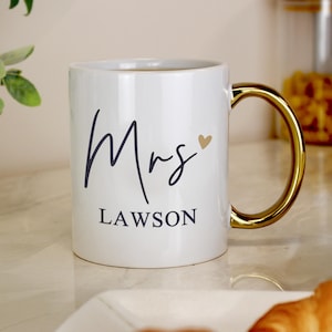 Pode incluir: Caneca de cerâmica branca com alça dourada. A caneca apresenta a palavra "Mrs" em fonte manuscrita, um coração dourado e o nome "Lawson" em fonte serifada. A caneca está cheia de um líquido castanho.