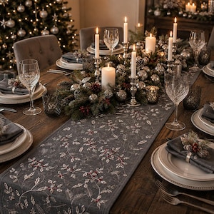 May include: A dark gray table runner featuring white embroidered holly leaves and red berries, set on a wooden dining table with white plates, gray napkins, silver napkin rings, crystal glassware, and a centerpiece of candles and ornaments.