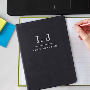 May include: A black notebook with a green spine. The front cover has the initials "LJ" in white with the name "LUKE JOHNSON" printed below in white.