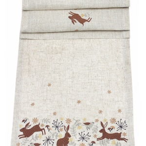 Può includere: Un runner da tavolo in lino beige con un ricamo di conigli marroni e uccelli in un campo di fiori. Il motivo è ripetuto sul bordo del runner.