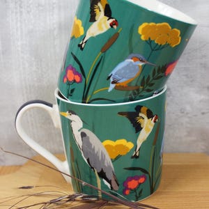 Könnte beinhalten: Zwei gestapelte Keramikbecher mit teal-farbenem Hintergrund und Vogelillustrationen. Die Becher haben einen weißen Griff und Rand, mit einem Design aus bunten Vögeln, Blumen und Schilf. Die Becher stehen auf einer Holzoberfläche.