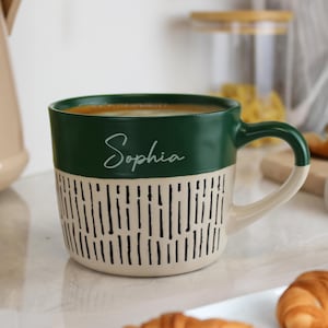 Könnte beinhalten: Eine Keramiktasse mit grünem Oberteil und cremefarbenem Boden mit einem schwarzen vertikalen Linienmuster. Das Wort "Sophia" ist in Kursivschrift auf dem grünen Abschnitt geschrieben. Die Tasse ist mit einem kaffeeähnlichen Getränk gefüllt.